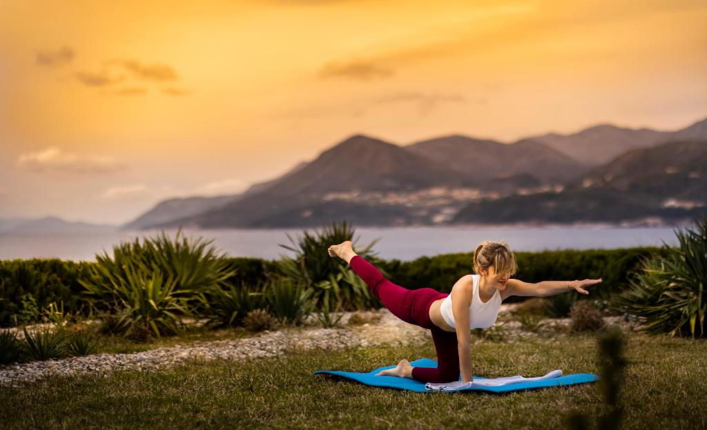Sieviete veic jogas pozas saulrietā, izmantojot jogas paklāju, ar kalniem un jūru fona.