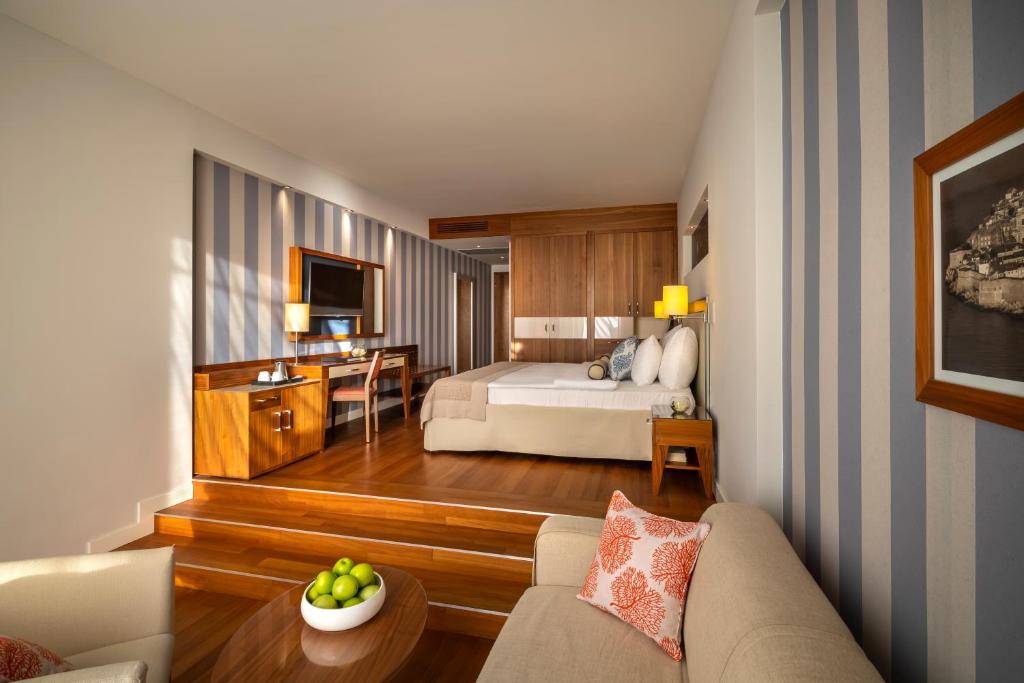 Mūsdienīgs viesnīcas numurs ar koka grīdu, gaisīgu interjeru un elegantām detaļām, piemēram, dīvānu un televizoru. Numurā redzams arī rakstāmgalds un augļu trauks ar zaļām āboliem.