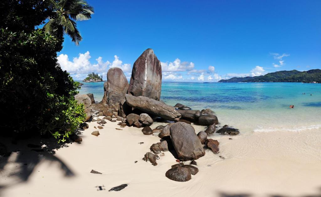 Skaists tropu pludmales skats ar lieliem akmeņiem un kristāldzidru ūdeni, kurā redzami cilvēki. Apkārt aug zaļi koki un zils debesis ar mākoņiem.