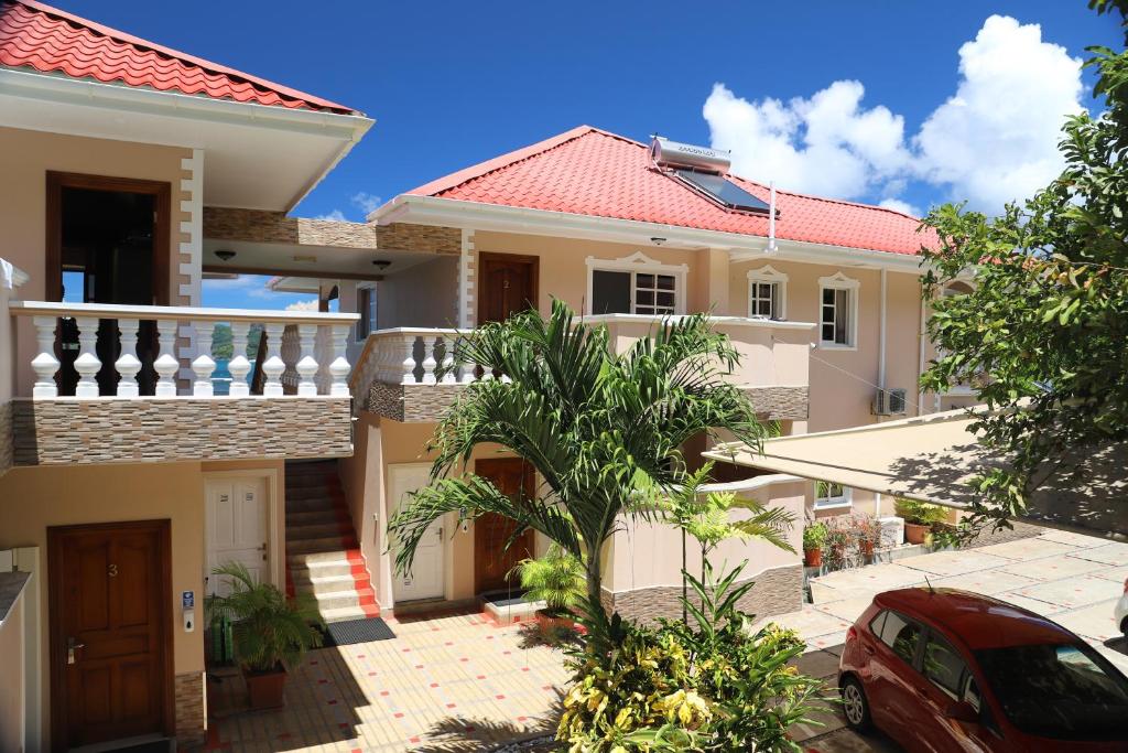 Mūsdienīga divstāvu māja ar sarkanu jumtu un balkonu, apkārt esošajiem zaļumiem un saulainu zilu debesu fonu. Iespēja redzēt autostāvvietu un augu dekorus.