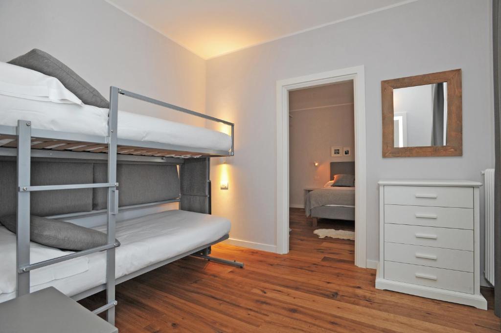 Moderns guļamistabas interjers ar divguļamo gultu un bunkura gultu, gaišiem toņiem un koka grīdu. Redzama atsevišķa telpa ar gultu un spoguli.