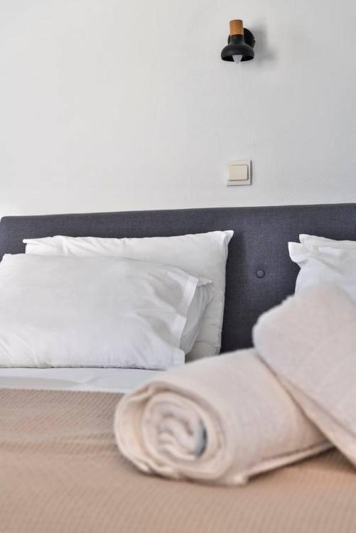 Mūsdienīgs guļamistabas interjers ar pelēku gultas galvu un baltām spilveniem, uz gultas sarullēts segu. Sienā piestiprināta stilīga lampiņa.