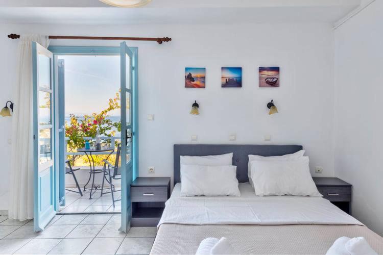 Mūsdienīgs guļamistabas interjers ar gaismu, kas ieplūst caur atvērtajām durvīm, un skatu uz terasi. Sienā ir izkārtoti gleznas, un gultā ir baltas spilveni un gultas veļa.