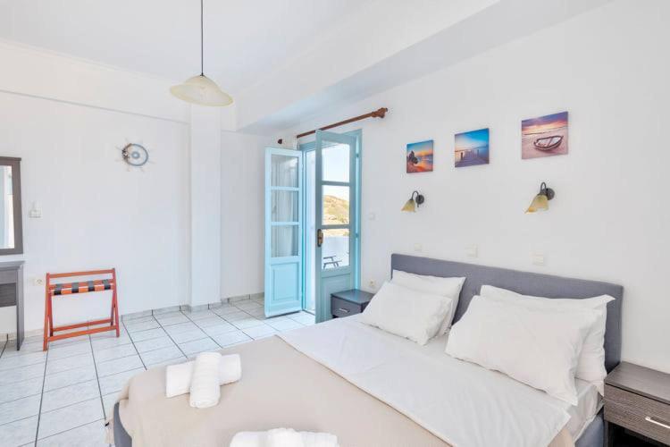 Gaiša un moderna guļamistaba ar divguļamo gultu, kas izrotāta ar siltām krāsām un piekļuvi balkonam caur zilo durvju atvērumu. Pie sienas ir nelieli attēli, bet telpā valda mierīga atmosfēra.