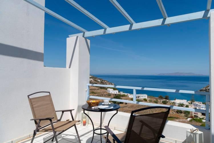 Balkons ar skatu uz jūru, divām krēslām un mazu galdiņu, uz kura atrodas kafijas krūze un uzkodas. Skats uz mierīgu jūru un kalnu ainavu.