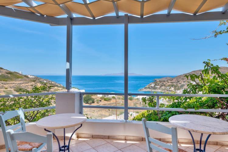 Skats no terases uz jūru, kur redzams gaišs zils debesis un kalnu ainava. Balti galdi un krēsli piedāvā ērtu vietu atpūtai.