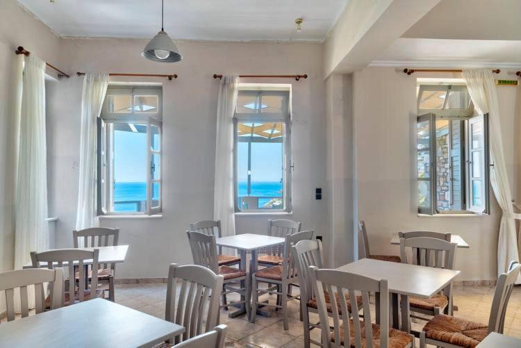 Gaiša kafejnīca ar baltām mēbelēm un skatu uz jūru caur atvērtajiem logiem. Interjers ir vienkāršs un mājīgs, radot relaksējošu atmosfēru.