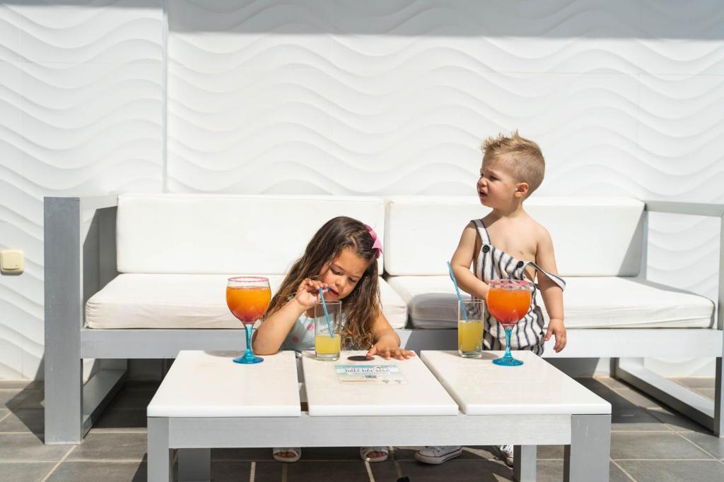 Divi bērni sēž pie baltas terases galda, baudot dzērienus no krāsainām glāzēm. Aiz viņiem redzama mūsdienīga, gaiša vide ar viļņveida sienu.