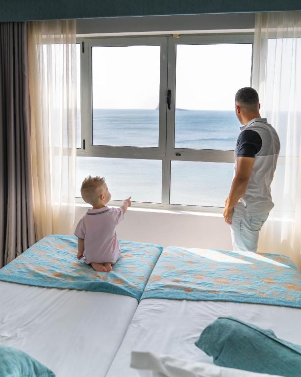Tēvs un dēls skatās pa logu uz jūru no viesnīcas numura. Bērns, sēžot uz gultas, rādīs uz kaut ko interesantu ārā, kamēr tēvs stāv blakus un vēro skatu.