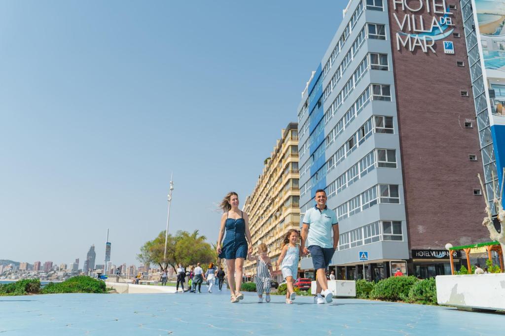 Ģimene pastaigā pa piejūras promenādi, ar viesnīcu "Hotel Villa del Mar" fonā. Skaists laiks un pilsētas ainava.