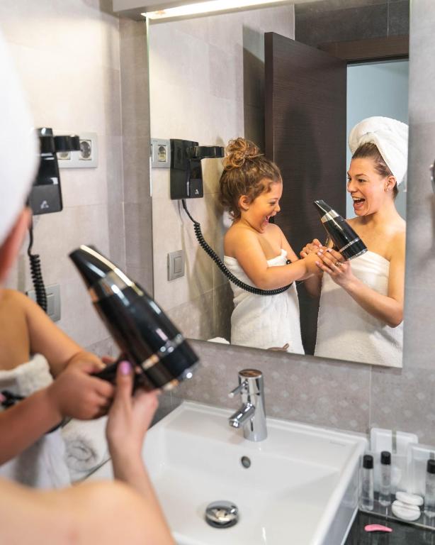 Mamma un meita jautrā brīdī vannas istabā, izmantojot matu fēnu, ar smaidiem un prieku.