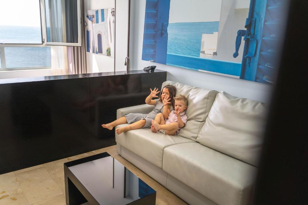 Divi bērni sēž uz pelēka dīvāna, priecīgi pozējot kamerai. Fons attēlo jūras skatu un gaišas, baltas ēkas.