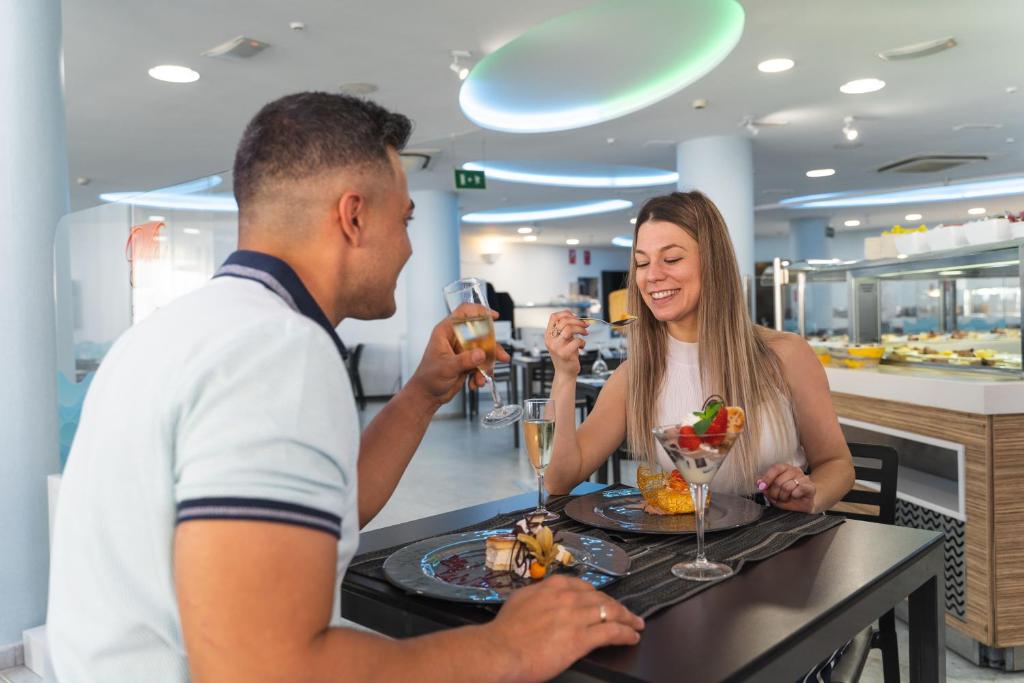 Romantisks brīdis, kur divi cilvēki bauda desertus un dzērienus modernā restorānā. Uz galda redzami gardi saldumi un kokteiļi, kas rada patīkamu atmosfēru.