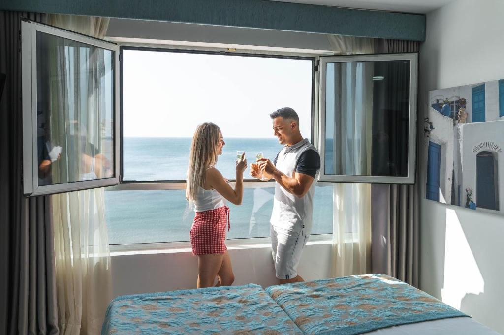 Pārī, kas svin savu laiku kopā, stāv pie atvērta loga ar skatu uz jūru, turēdami dzērienus rokās. Tiek radīta romantiska atmosfēra, baudot brīnišķīgu skatu un katru brīdi.