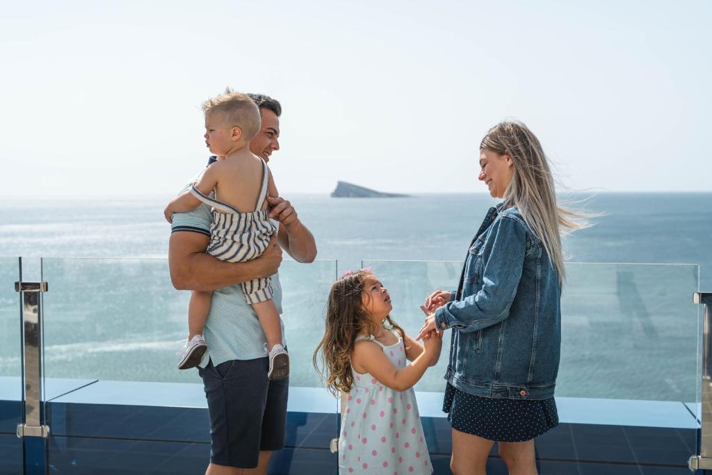 Ģimene, kas stāv uz terases ar skatu uz jūru, tēvs tur bērnu rokās, kamēr meitene sarunājas ar māti. Fons ir jūra un neliela sala.