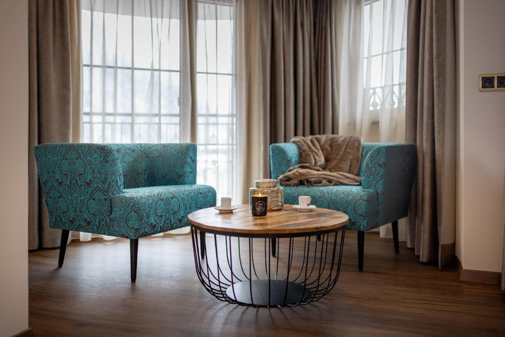 Mūsdienīga dzīvojamā telpa ar divām elegantiem zaļiem krēsliem un koka galdu, uz kura atrodas sveces un kafijas tasītes. Labi apgaismota ar logiem, kas rada mājīgu atmosfēru.