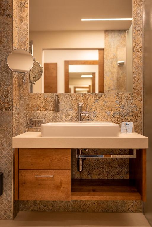 Mūsdienīgas vannas istabas dizains ar elegantu izlietni un spoguli, pie kura ir redzami atkārtoti atstarojoši elementi. Siltas koka akcenti un grezns apdare veido patīkamu atmosfēru.