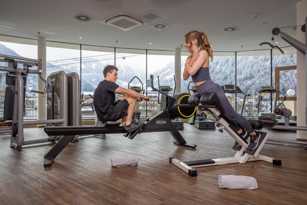 Divi cilvēki sporto modernā fitnesa zālē ar skatu uz kalniem, izmantojot kardio un spēka treniņu aprīkojumu.