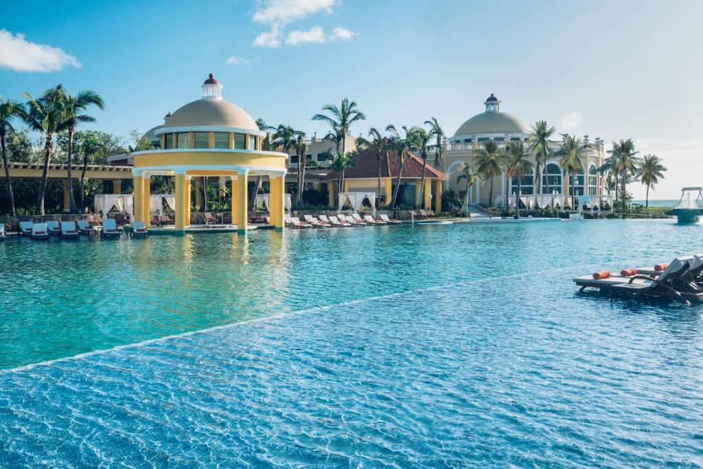 Luksusa kūrorts ar plašu baseinu un palmām, kas ieskauj ēkas ar skaistu arhitektūru un saulainu debesi.