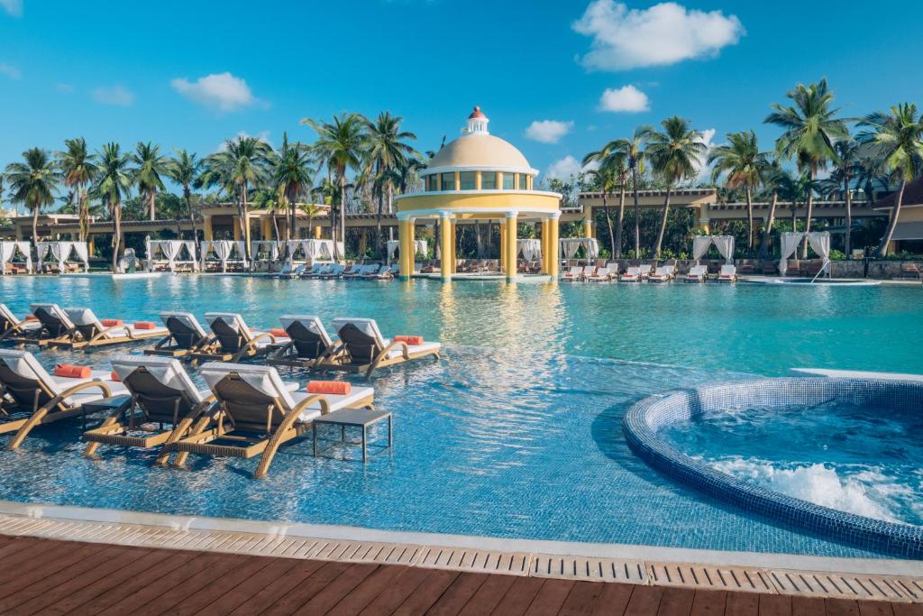 Luksusīgs kūrorta baseins ar saulessargiem un palmām, kas rada atpūtas atmosfēru. Ūdens ir dzidrs un mirdzošs, savukārt apkārtējā vide ir eleganta un relaksējoša.