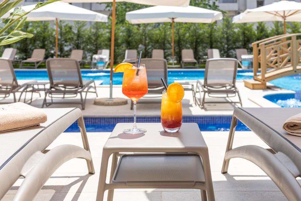 Divi kokteiļi atpūtas zonā blakus baseinam, ar sauļošanās krēsliem un saulessargiem fonā. Ideāla vieta relaksācijai un atpūtai.