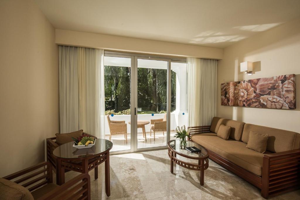 Mūsdienīga viesnīcas viesistaba ar koka mēbelēm, apaļu galdu un skatu uz dārzu caur lieliem logiem. Telpa ir gaiša un gaumīga, ar ērtu dīvānu un dekoratīvu mākslu uz sienas.