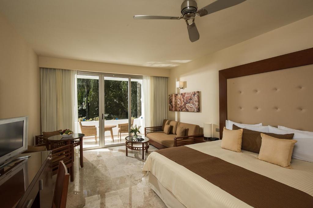 Mūsdienīga viesnīcas numura iekārtojums ar ērtu gultu, dīvānu un lieliem logiem, kas ved uz terasi. Numurā ir televizors un galda zona, radot relaksējošu atmosfēru.