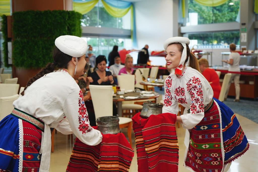 Divas sievietes tradicionālās tautas tērpos, nēsājot traukus, piedalās kultūras pasākumā viesnīcā. Fonā redzamas apmeklētāju grupas, kas bauda maltīti un sarunas.