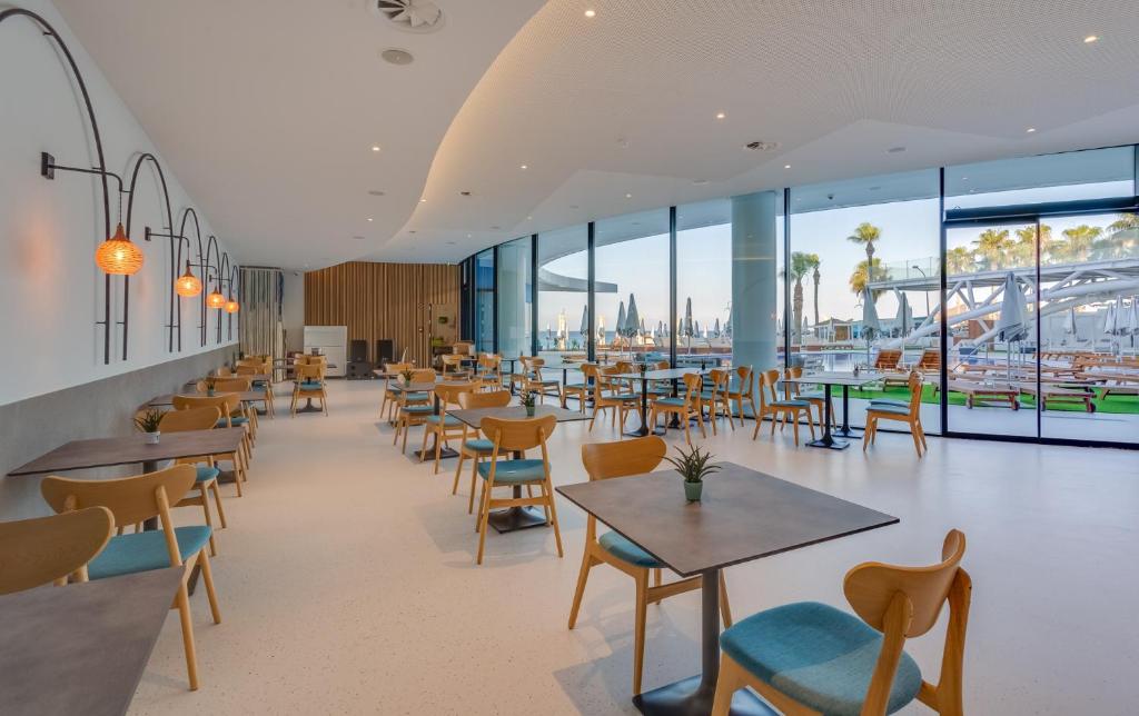 Mūsdienīgs restorāns ar gaišu interjeru, kurā ir koka mēbeles un lieli logi, kas paver skatu uz āra teritoriju ar palmām.