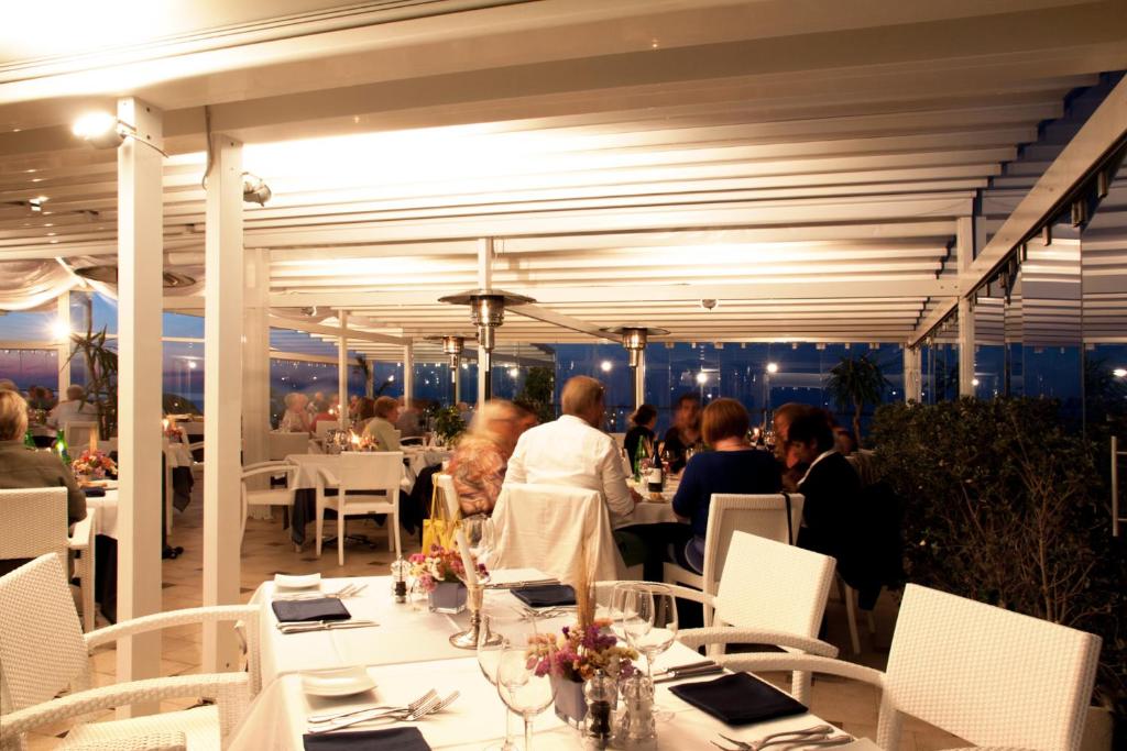 Elegants restorāns ar moderna dizaina interjeru, kur viesi bauda maltītes vakarā. Paveras skats uz apgaismotu pilsētu caur stikla logiem.