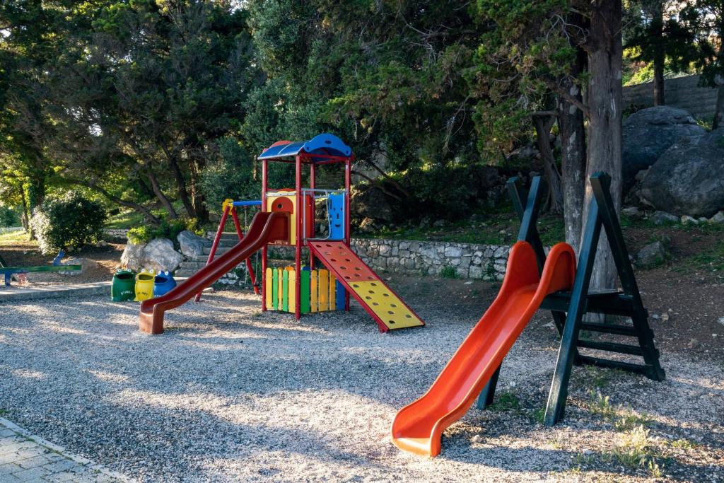 Krāsains bērnu rotaļu laukums ar divām slidkalniņām un rotaļu struktūru, kas ielikta dabiskā vidē. Apkārt ir zaļumi un akmeņi.