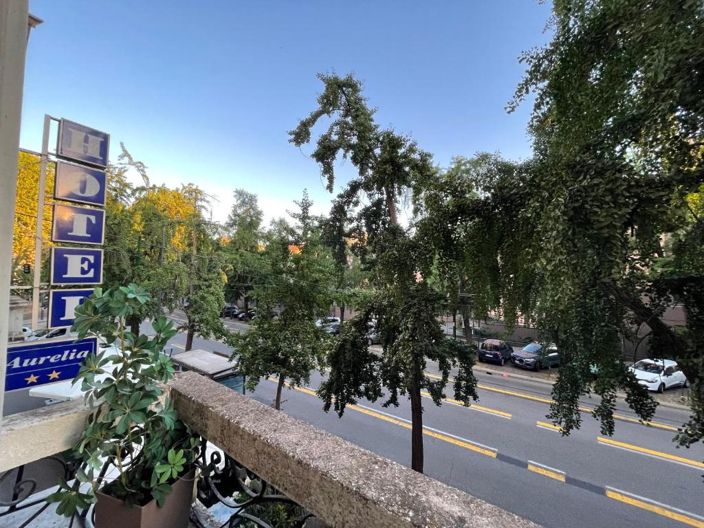 Skats no viesnīcas balkona ar kokiem un ielu, kurā redzamas automašīnas. Viesnīcas zīme "Aurelia" ir redzama kreisajā pusē.