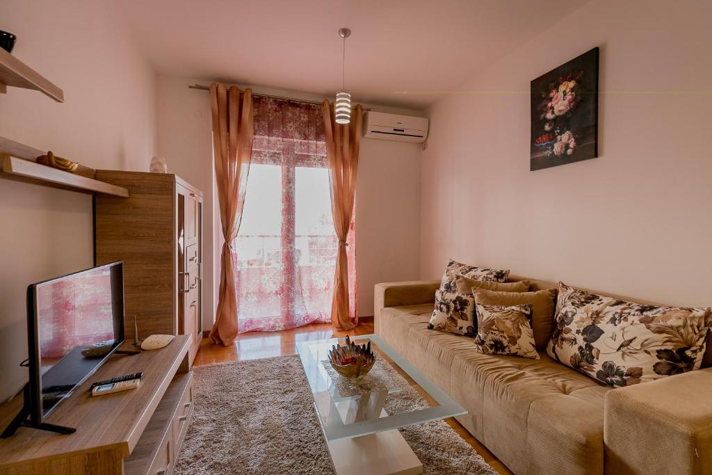 Mūsdienīgs viesistabas interjers ar dīvānu, televizoru un gaisīgām aizkariem, kas atveras uz balkonu. Uz galda izvietotas dekoratīvas detaļas, radot mājīgu atmosfēru.