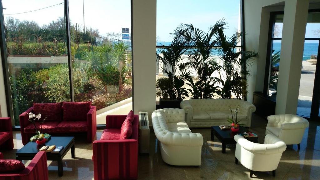 Mūsdienīgs viesnīcas vestibilis ar komfortablām sarkanām un baltām mēbelēm, caur lielajiem logiem redzama ainava ar zaļumiem un jūras skatu.