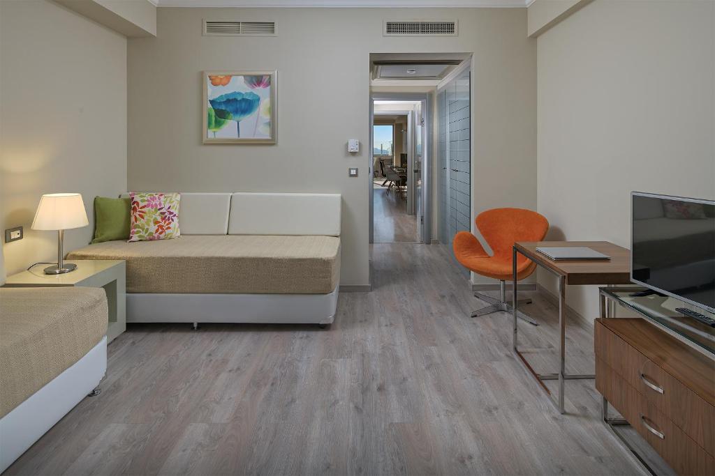 Mūsdienīga viesnīcas numura interjers ar gaišām krāsām, ērtām mēbelēm un panorāmas skatu uz koridoru. Telpā ir dīvāns, oranža krēsla un televizors.