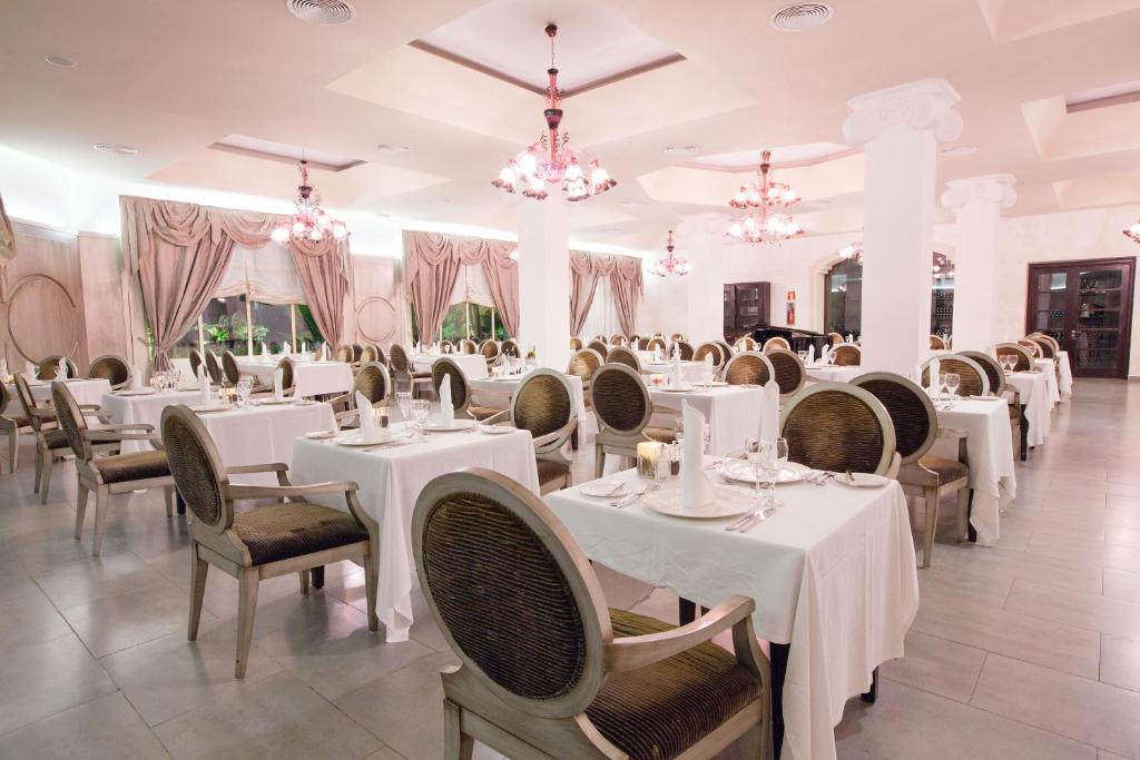 Elegants restorāns ar baltiem galdautiem un greznām krēslām, apgaismots ar rozā lustru gaismu. Interjers ietver eleganti drapētas aizkarus un mūsdienīgu dizainu, radot viesmīlīgu atmosfēru.