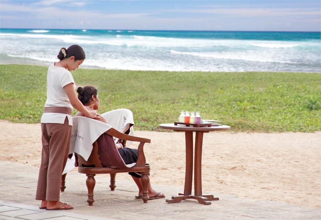 Piedāvājot relaksējošu masāžu pludmalē, šis attēls parāda, kā profesionāls masieris sniedz pakalpojumu viesim, kamēr fona ainava ietver skaistu jūru un zāles laukumu.