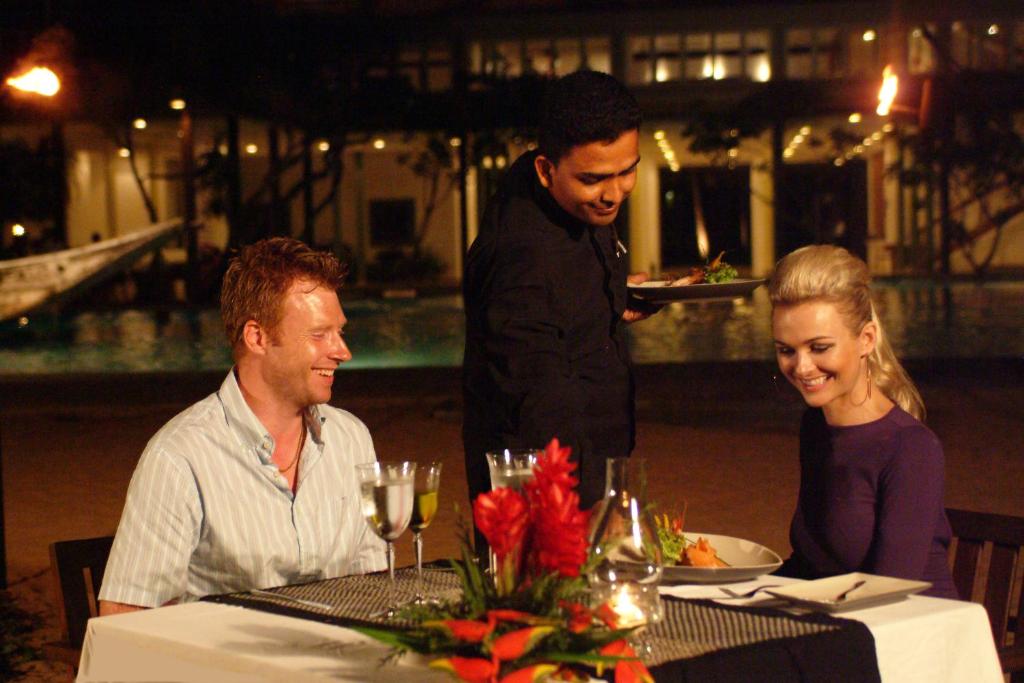 Romantiska vakariņas pie baseina, kur viesmīlis pasniedz ēdienu diviem apmierinātiem viesiem. Galda dekors ir krāšņs ar ziediem un dzērieniem.