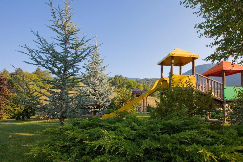 Bērnu rotaļu laukums ar dzelteno slidkalniņu, ko ieskauj zaļi krūmi un egļu koki. Fons ar kalnu ainavu un skaidru debesu.