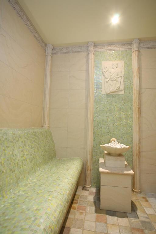 Relaksējoša pirts telpa ar zaļganu mozaīkas soli un elegantu strūklaku centrā. Sienas rotātas ar izsmalcinātu reljefu un gaišiem akmeņiem.