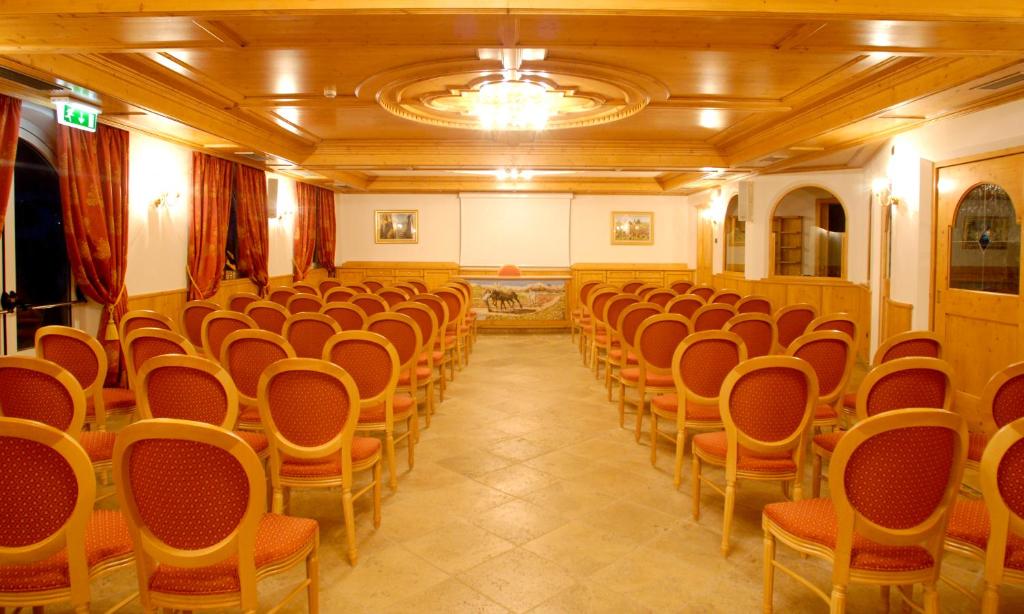 Konferenču telpa ar koka apdari un sarkaniem krēsliem, organizēta rindu veidā. Telpā ir gaismas avoti un logi ar aizkariem, piedāvājot siltu un viesmīlīgu atmosfēru.