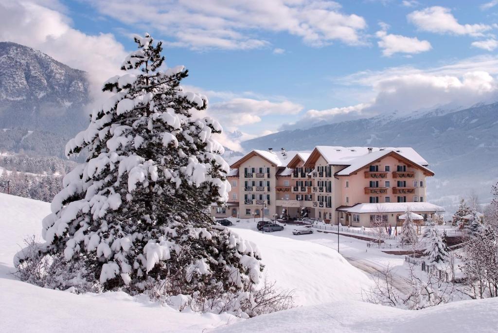 Sniegotā ainava ar viesnīcu fona, ko ieskauj kalni un sniegots priedes koks priekšplānā.