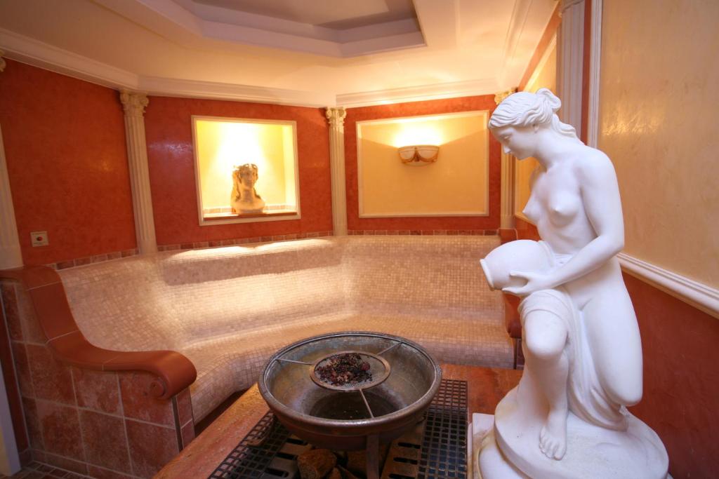 Elegants wellness telpas interjers ar baltu skulptūru un siltām krāsām, radot relaksējošu atmosfēru. Centrā ir apaļš baseins, ap kuru izvietotas sēdvietas un dekoratīvi elementi.