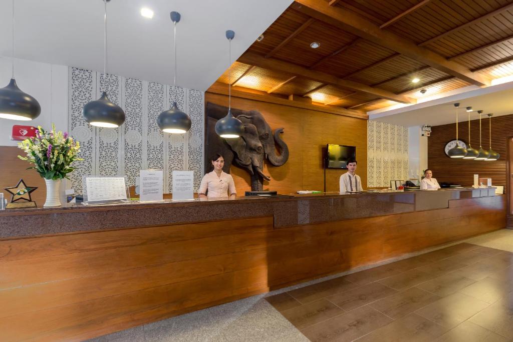 Recepcija ar modernu interjeru, kurā ir koka elementi un dekoratīvs zilonis uz sienas, apkalpo viesus. Pie letes strādā trīs darbinieki, un uz galda ir ziedi un informācijas lapas.