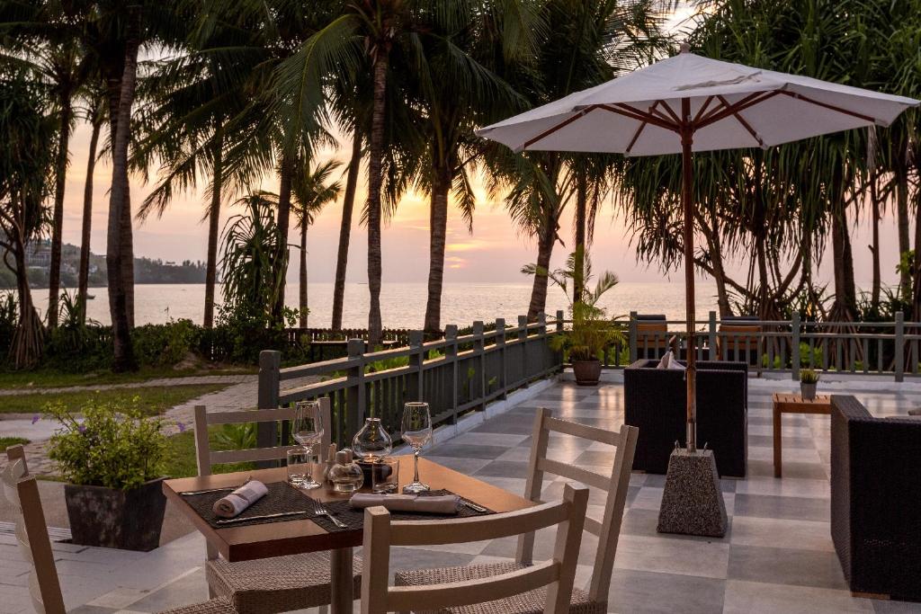 Elegants restorāns ar skatu uz jūru, apzaļumots ar palmām un terasi, kurā ir galds ar traukiem un šampanieša glāzēm. Saule noriet aiz horizonta, radot mierīgu atmosfēru.