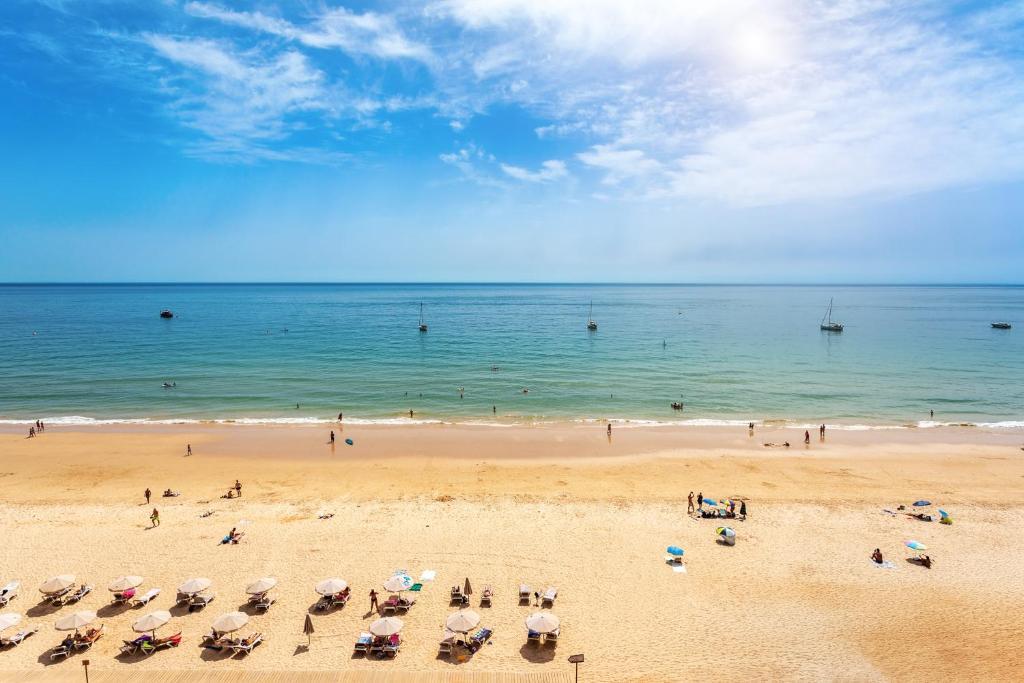 Skaists pludmales skats ar smilšu krastu, apmeklētājiem un kuģiem jūrā. Zilā debess ar vieglām mākoņu svītrām rada mierīgu atmosfēru.