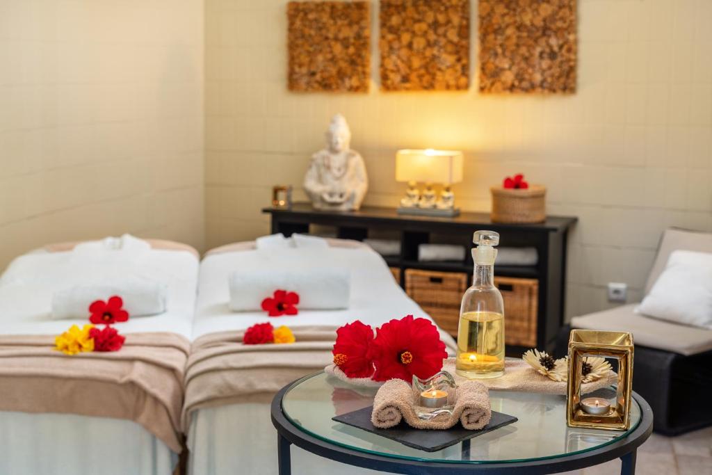 Relaksējoša spa telpa ar divām masāžas galdiem, dekorēta ar ziediem un mākslas priekšmetiem, radot mierīgu atmosfēru. Uz galda atrodas aromātiska eļļa un svece, kas veicina relaksāciju.