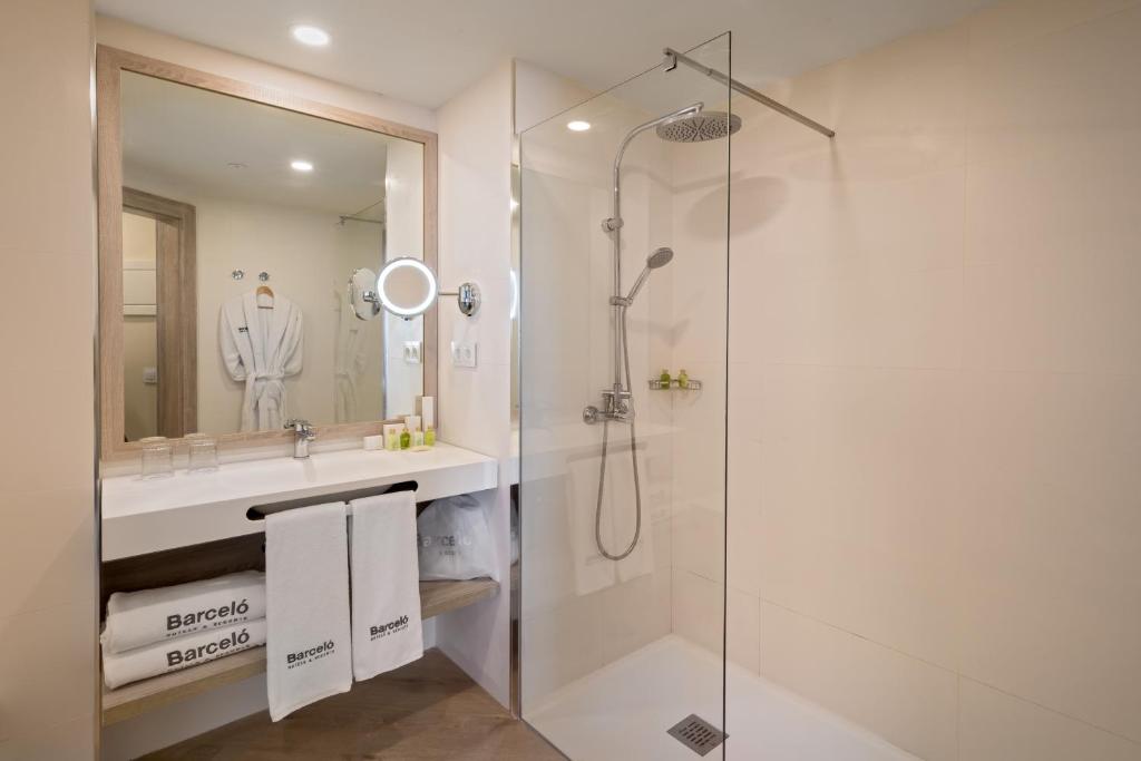 Mūsdienīga un stilīga vannas istaba ar stikla dušu, lielu spoguli un ērtu izlietni. Redzami balti dvieļi ar "Barceló" logo un dažādi higiēnas produkti.