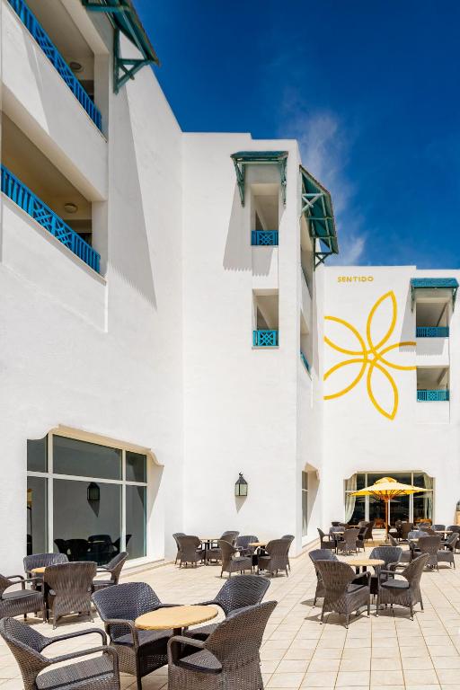 Balta viesnīca ar zilu balkonu un dārza terasi, kurā redzami vīna galdi un krēsli zem saules. Uz ēkas sienas ir dzeltena ziedu ilustrācija.