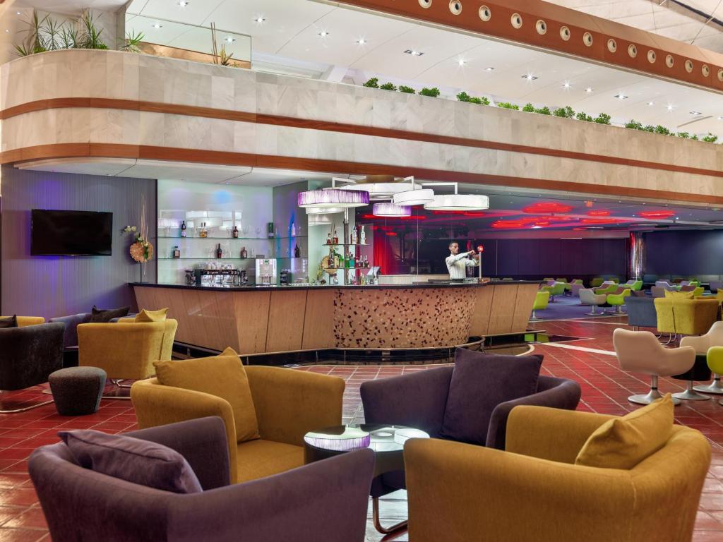 Mūsdienīgs bārs ar krāsainiem sēdekļiem un stilīgu apgaismojumu, kas rada relaksējošu atmosfēru. Interjerā ir redzams arī bāra galds un dzērienu plaukti.